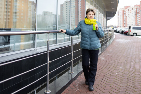 Mature woman in warm jacket and bright scarf holding reusable coffee cup on urban balcony, thoughtful look, city background.