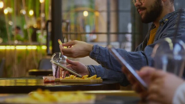 Young man with galsses and beard snaking on french fries with ketchup while scrolling social media on smartphone at table in modern fast food cafe