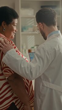Vertical rear view medium shot of Middle Eastern doctor showing blood sugar tracking application on smartphone to senior Black woman during healthcare visit