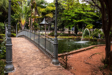 View of a classic metal bridge crossing a pond with fountains at a public garden in the city of Amparo, Sao Paulo state, Brazil.