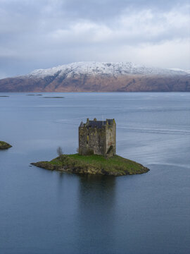 Aerial view of Castle Stalker on a small tidal islet surrounded by the calm waters of Loch Laich with snow-capped mountains under a cloudy sky Appin, Scotland, United Kingdom.