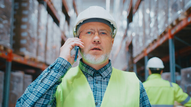 Confident mature man with short gray beard wearing helmet and vest. Speaking on smartphone in warehouse. Looking ahead. Resolving issue or leading conversation during supply operation.