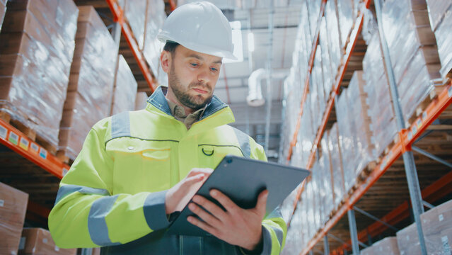 Bearded Caucasian man in high-visibility jacket and helmet using digital tablet while standing near tall racks in warehouse. Reviewing stock data or updating inventory information during inspection.