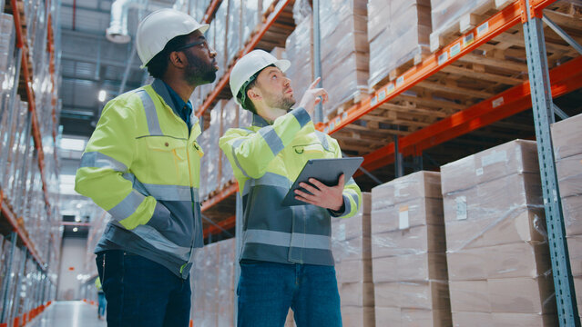 Warehouse workers in helmets and reflective jackets discussing logistics in front of packed shelves. Male pointing upward. Partner watching direction. Planning storage layout or stock placement.