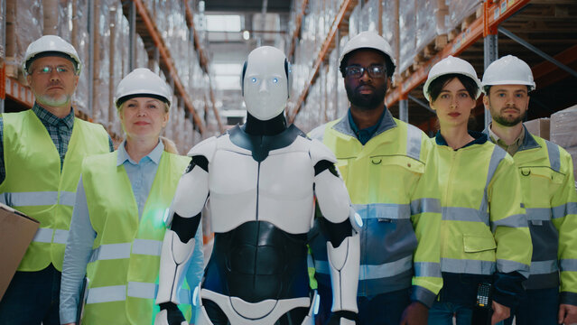 Group of warehouse workers standing beside humanoid robot in aisle lined with pallets. People wearing safety vests and hard hats. Posing proudly with scanning device and boxes in logistics facility.