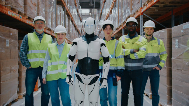 Team of warehouse workers posing with humanoid robot in distribution center aisle. Wearing hard hats and reflective vests Standing confidently beside robotic coworker in middle of stacked pallets.
