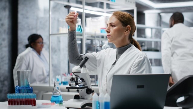 Scientist in lab pipetting blue liquid into test tube to analyze it. Laboratory worker transferring chemical sample to prepare for immediate spectroscopic analysis, camera B
