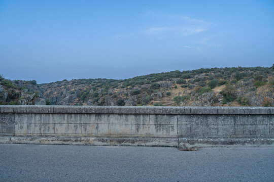 A weathered stone bridge parapet overlooks a paved road, with rocky hills and scattered vegetation under a cloudy blue sky.