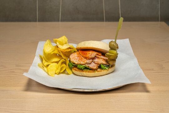 A delicious sandwich on a roll with pork loin steaks, potato chips in a bag, and a skewer.