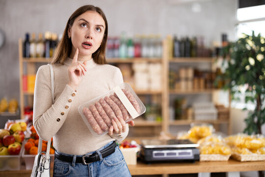 Young woman in store examines raw bbg sausage, chooses raw meat for dinner. Buyer examines information on label, examines condition of food.