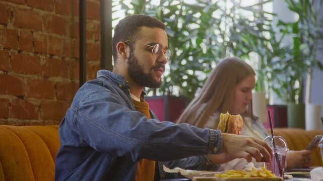 Young bearded man in glasses pouring ketchup and taking bite of tasty burger at table in modern cafe