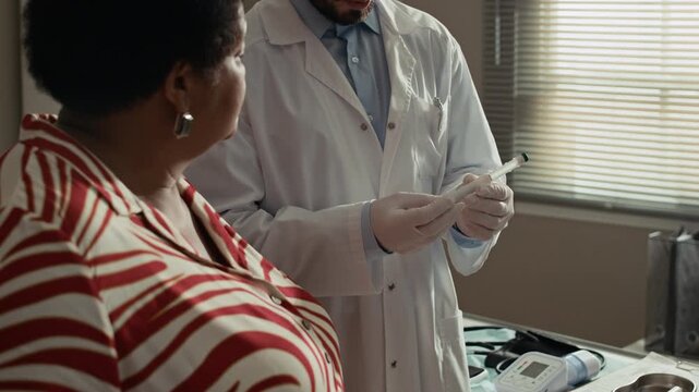 Medium shot of Middle Eastern male doctor administering insulin injection pen to senior Black female patient during diabetes appointment in modern medical office