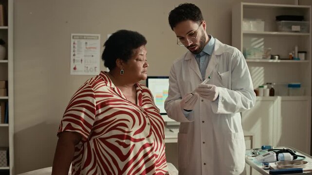 Medium shot of Middle Eastern physician using insulin injection pen on older Black female patient during diabetes treatment consultation in modern medical office
