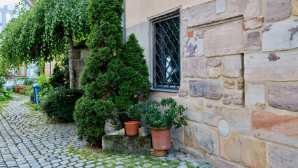 Obraz premium Cozy courtyard of an old German house with a rustic sandstone wall, small window with iron bars, green thuja trees, and a cobblestone pathway.