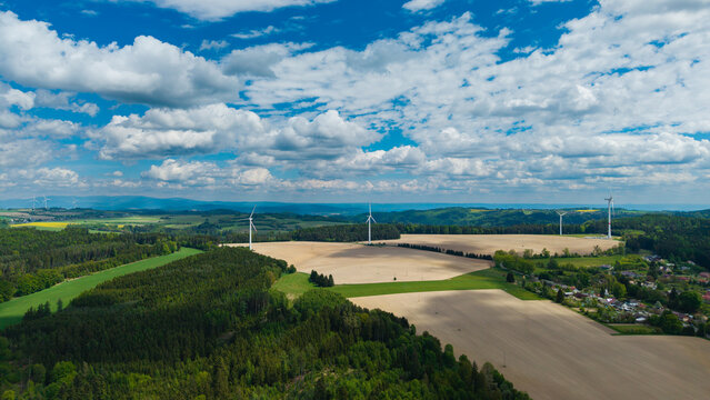 Aerial view of wind turbines standing in open fields surrounded by dense green forests under a blue sky with white clouds near Zipotin Gruna, Pardubicky kraj, Czechia.