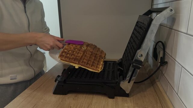 Cook removes freshly baked waffles from waffle maker with spatula placing them onto plate. 