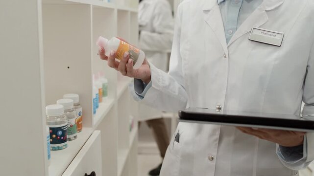 Tilt up detail shot of Caucasian young woman wearing white coat and using tablet while doing stock inventory in pharmacy storage room, copy space