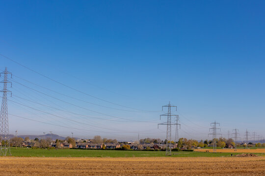High-voltage power lines stretch across the rural landscape of Vis&eacute;, Belgium, A clear blue sky meets fertile farmland and a quiet village, illustrating the intersection of energy and countryside.
