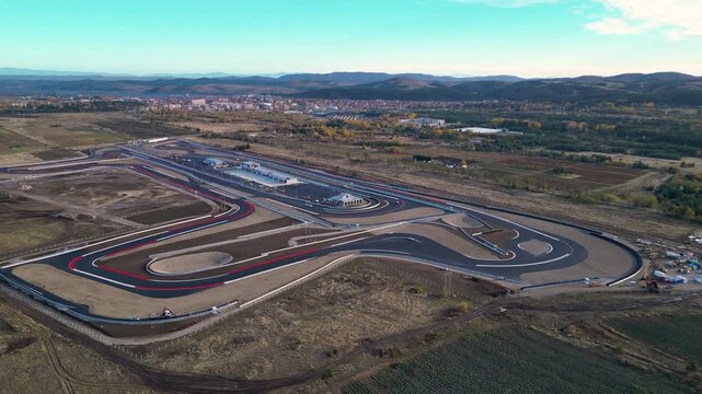 Aerial view of a modern motorsport racetrack with sweeping curves, pit buildings, and wide runoff areas set in an open rural landscape near a town and distant hills