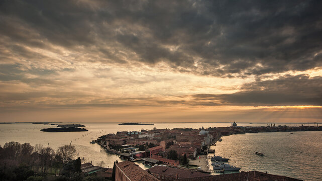 Aerial view of the historic rooftops and islands of the Venetian Lagoon under a dramatic cloudy sky with sun rays at sunset Venice, Veneto, Italy.