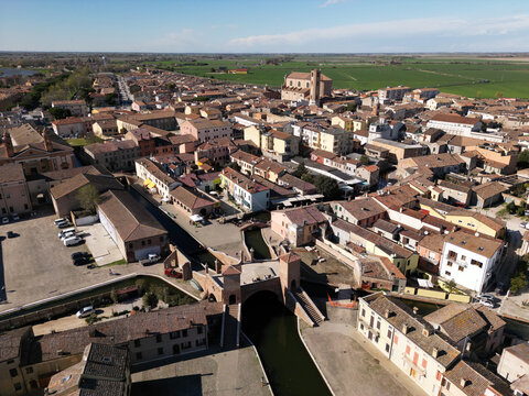 Aerial view of the historic Trepponti bridge and scenic canals surrounded by traditional terracotta rooftops and green fields in Comacchio, Emilia-Romagna, Italy.