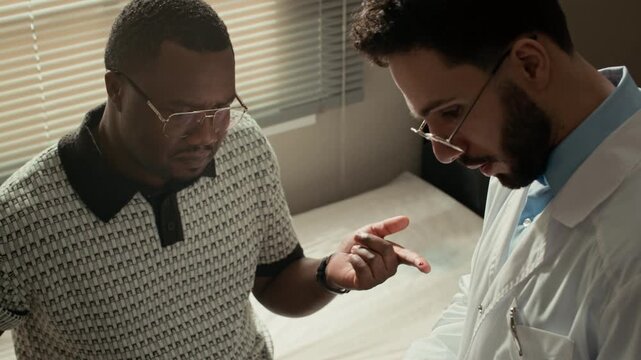 Tilt up of Middle Eastern male doctor performing finger blood sugar test with glucometer on Black male patient during diabetes consultation in clinic