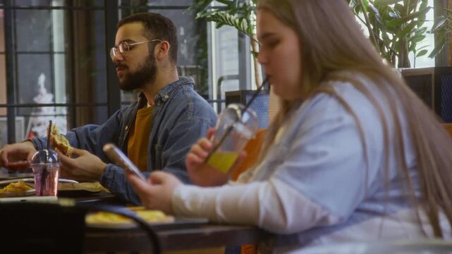 Young woman sipping drink and scrolling social media on smartphone while biracial man enjoying burger at separate table in modern fast food restaurant