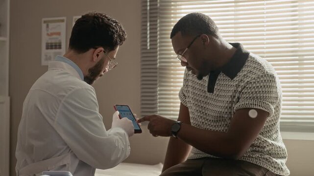 Side view medium shot of Black male patient looking at smartphone screen with glucose monitoring app while Middle Eastern male doctor providing guidance during healthcare visit