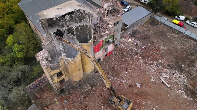 High-angle drone shot of a partially collapsed structure surrounded by forest, with an excavator arm reaching across the remains of the building and water being used to suppress dust during demolition