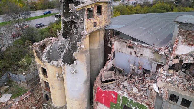 Aerial drone footage of a partially demolished industrial building featuring large concrete silos, collapsed masonry walls, and piles of construction debris. The scene documents urban redevelopment, i