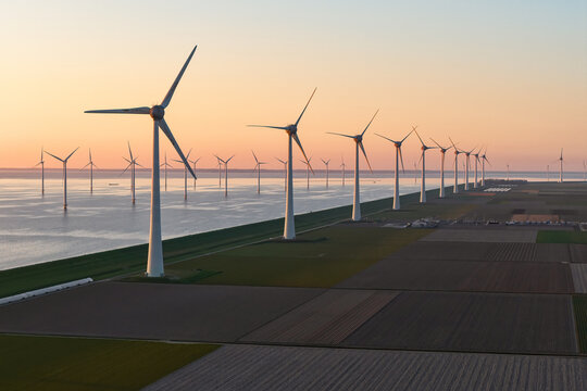 Aerial view of a row of modern wind turbines standing along a coastal dike between agricultural fields and the sea during a golden sunset in the Netherlands.