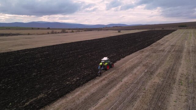 Aerial drone footage following a modern tractor pulling a mounted plow while turning soil across a large agricultural field. The clip highlights seasonal land preparation, mechanized farming, soil cul