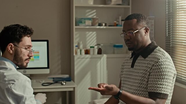 Arc waist up shot of Middle Eastern male doctor wearing white coat, gloves and glasses using blood glucose meter on Black male patient finger during routine diabetic health examination in clinic