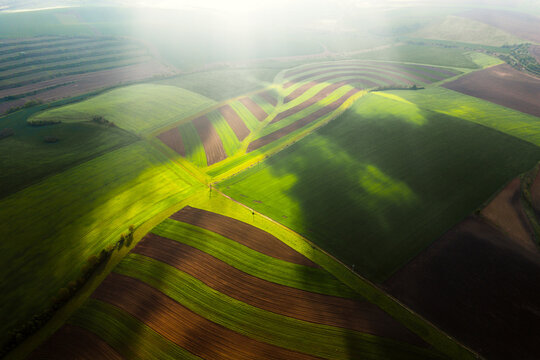 Aerial view of rolling agricultural fields with green crops and brown soil under dramatic sunlight and cloud shadows in Klobouky u Brna, Jihomoravsky kraj, Czechia.