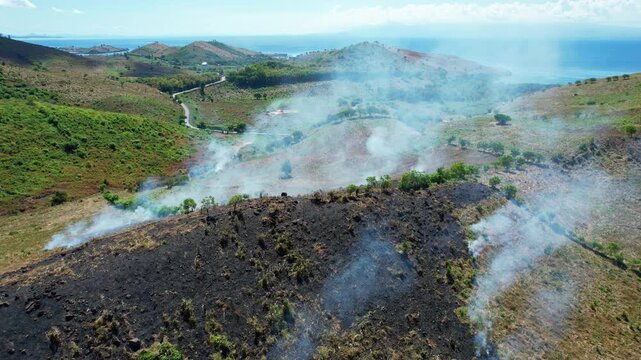 Aerial drone footage of a burning corn crop hill, with many smokes, made to regenerate the lands, in Kwangko area, Dompu regency, Sumbawa island, Indonesia, with hills and blue sea behind