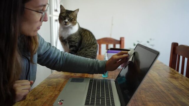 Woman cleaning laptop screen with cat watching