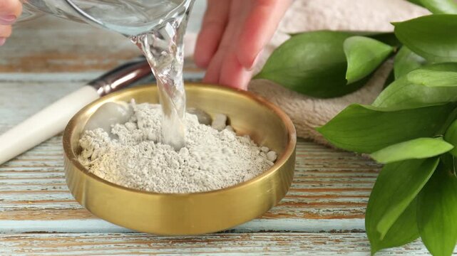 Making clay paste for hair and skin care. Woman pouring water into bowl with clay powder and stirring it with makeup brush at color wooden table, closeup. Mud therapy