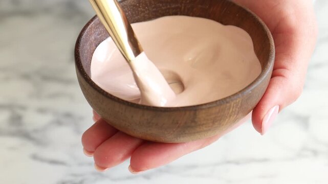 Woman stirring clay paste for hair and skin care with makeup brush at white marble table, closeup. Mud therapy