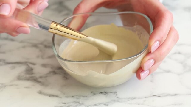 Woman taking natural clay paste for facial and body care from bowl with makeup brush and applying it onto her hand at white marble table, closeup. Mud therapy