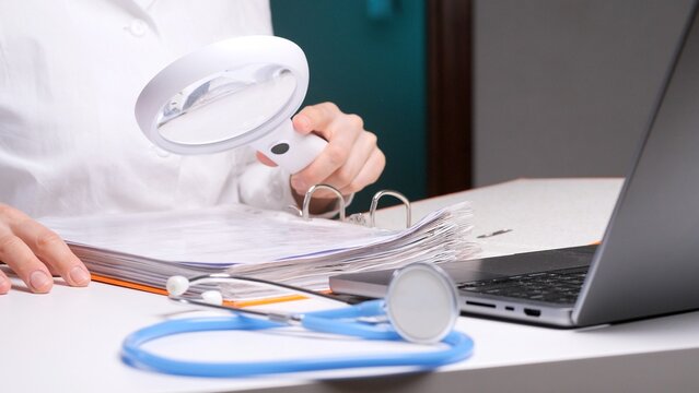 Doctor's hands carefully examining patient health records with a magnifying glass, verifying details on medical history and diagnostic reports while a stethoscope and laptop are on the desk