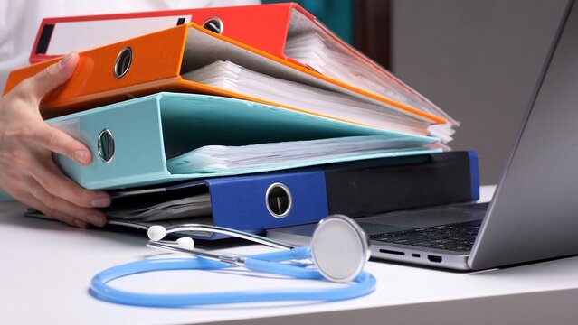 Healthcare professional balancing a heavy stack of patient medical records and files, symbolizing overwhelming bureaucracy and administration, while a stethoscope and laptop sit on the desk