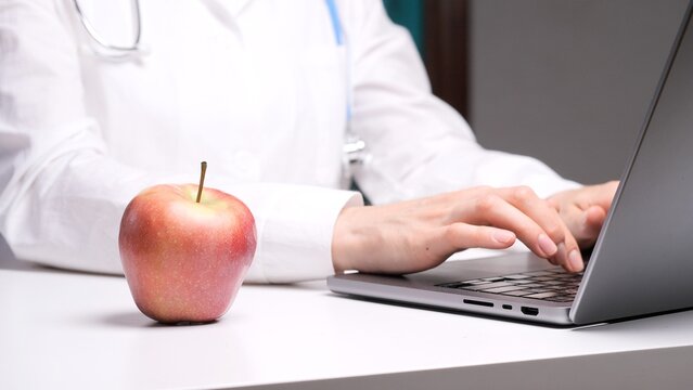 Medical professional nutritionist at a desk with apple, typing on laptop to provide telemedicine and online dietary advice for patient wellness and healthy eating guidance