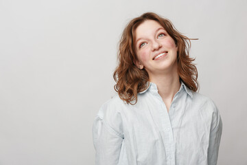 Obraz premium Young woman with red hair and light blue shirt looks up smiling on grey background. Concept of happiness, hope, casual style, and natural beauty in studio portrait.