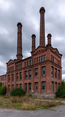 Fototapeta premium Brickwork's Testament: An aged brick industrial building, a relic of bygone eras, stands tall beneath a cloudy sky, showcasing the enduring strength of architecture.
