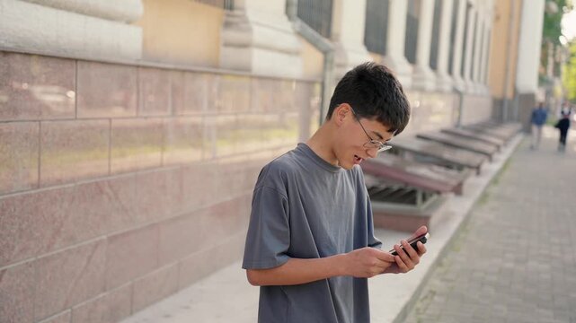 Expressive young person reacting emotionally to smartphone message with gestures outdoors. Frustrated boy expressing negative emotions. Authentic lifestyle, communication and emotions concept.