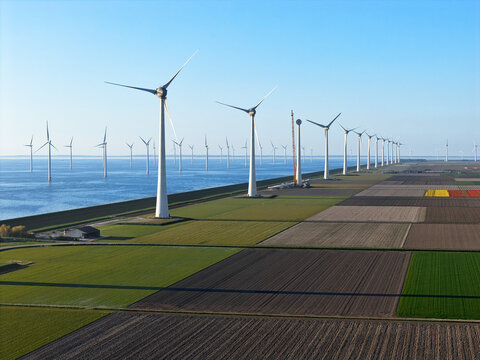 Aerial view of a row of wind turbines standing along a coastline next to colorful agricultural fields and the sea under a clear blue sky Netherlands.