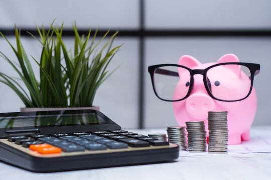Pink piggy bank wearing eyeglasses and calculator, stacks of coins and potted plant on financial desk. Concept savings, budgeting, accounting and financial planning for business and investment themes.