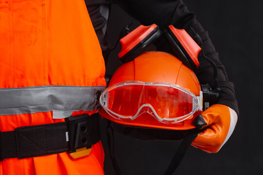 Worker in orange hi-vis vest holding hard hat with safety goggles and earmuffs. Concept for personal protective equipment, workplace safety and construction industry.