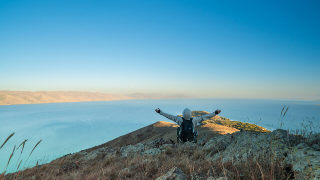 A hiker standing on a mountain overlooking Lake Sevan, Armenia, with a backpack, admiring the breathtaking landscape of water and distant hills.