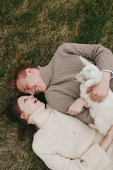 Fototapeta premium Smiling couple lying on grass with a curious goat kid, embracing and laughing in warm natural light. Cozy sweaters, outdoor vibe, intimate lifestyle moment with playful animal companionship.